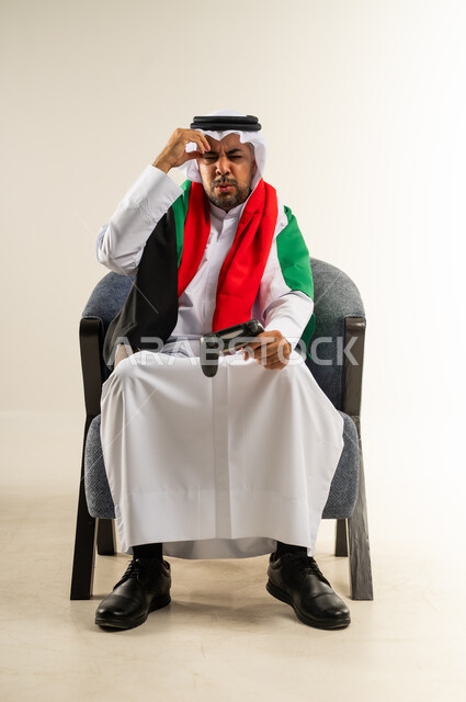 التسلية والترفيه بالفيديو جيمز في وقت الفراغ، إيماءات الخسارة وخيبة الأمل، استخدام الألعاب الإلكترونية والأجهزة الحديثة، بورتريه لرجل عربي خليجي كويتي يرتدي الثوب التقليدي وعلم الكويت على كتفه يمسك عصا التحكم يضع يده على رأسه بتعابير الحسرة والحزن، خلفية بيضاء