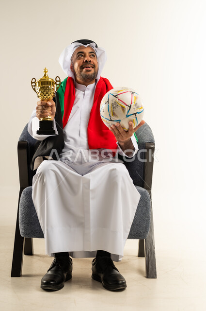Cheering for the favorite team, joy for the national team's victory, gestures of happiness and joy, portrait of a Kuwaiti Gulf Arab man wearing traditional thobe sitting on a comfortable chair holding a football and the golden World Cup in his hands looking at something above, passion for following the matches, white background