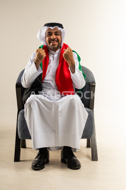 إيماءات السعادة والفرح، بورتريه لرجل عربي خليجي كويتي يرتدي الثوب التقليدي يجلس على كرسي مريح يضع علم الكويت على كتفيه ينظر إلى الكاميرا يرفع يديه بتعابير الانتصار، السرور لفوز المنتخب الوطني، تشجيع الفريق المفضل، الشغف بمتابعة مباريات كرة القدم، خلفية بيضاء