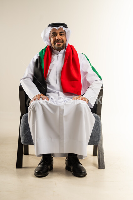 السرور لفوز المنتخب الوطني، إيماءات السعادة والفرح، بورتريه لرجل عربي خليجي كويتي يرتدي الثوب التقليدي يجلس على كرسي مريح يضع علم الكويت على كتفيه ينظر إلى الكاميرا، تشجيع الفريق المفضل، الشغف بمتابعة مباريات كرة القدم، خلفية بيضاء