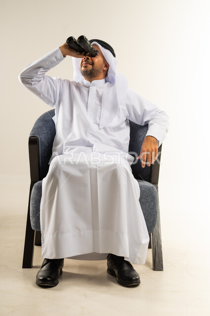 رؤية الأشياء البعيدة وتقريبها, حب الاستكشاف و المغامرة, بورتريه لرجل عربي خليجي كويتي يرتدي الثوب التقليدي يجلس على كرسي مريح ينظر من خلال منظار ثنائي العينين إلى شيئ ما, إيماءات الاستمتاع والسرور, خلفية بيضاء