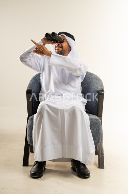 إيماءات الاستمتاع والسرور, رؤية الأشياء البعيدة وتقريبها, حب الاستكشاف و المغامرة, بورتريه لرجل عربي خليجي كويتي يرتدي الثوب التقليدي يجلس على كرسي مريح ينظر من خلال منظار ثنائي العينين يشير بإصبع السبابة إلى شيئ ما, خلفية بيضاء