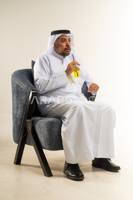 إيماءات الذهول والصدمة, محاولة التخفيف من حرارة الصيف بالمشروب المنعش، الاستمتاع بالمشروبات المفيدة اللذيذة، بورتريه لرجل عربي خليجي كويتي يرتدي الثوب التقليدي يجلس على كرسي مريح ويستمتع بشرب العصير الطبيعي البارد, خلفية بيضاء
