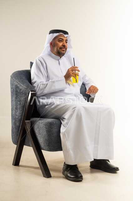 محاولة التخفيف من حرارة الصيف بالمشروب المنعش، الاستمتاع بالمشروبات المفيدة اللذيذة، بورتريه لرجل عربي خليجي كويتي يرتدي الثوب التقليدي يجلس على كرسي مريح ويستمتع بشرب العصير الطبيعي البارد, خلفية بيضاء
