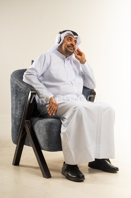 الاندماج والتفاعل مع الأنغام الموسيقية، مواهب غنائية فنية، بورتريه لرجل عربي خليجي كويتي يرتدي الثوب التقليدي يجلس على كرسي مريح يستمع إلى الموسيقى من خلال سماعة الرأس، الانصات والاستماع للأغاني باستخدام الهيدفون، خلفية بيضاء