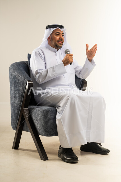 تعابير الاندماج والتفاعل مع الموسيقا، تسجيل مقطع صوتي غنائي، بورتريه لرجل عربي خليجي كويتي يرتدي الثوب التقليدي يجلس على كرسي مريح يمسك بيده المايكروفون يغني بإيماءات الحماس والسرور، تقديم العروض الصوتية، ممارسة موهبة الغناء، خلفية بيضاء