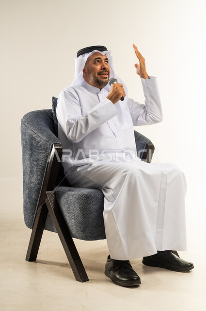 ممارسة موهبة الغناء، تسجيل مقطع صوتي غنائي، بورتريه لرجل عربي خليجي كويتي يرتدي الثوب التقليدي يجلس على كرسي مريح يمسك بيده المايكروفون يغني بإيماءات الحماس والسرور، تقديم العروض الصوتية، تعابير الاندماج والتفاعل مع الموسيقا، خلفية بيضاء
