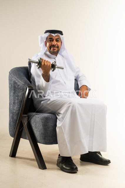 تقوية عضلات الجسم و بنائها ،المحافظة على الصحة واللياقة الجسدية والرشاقة البدنية، بورتريه لسيد عربي خليجي كويتي يرتدي الثوب التقليدي يجلس على الكرسي المريح يحمل بيده الأوزان الحديدية الثقيلة، ممارسة التمارين الرياضية ورفع الأثقال مع الدمبل، خلفية بيضاء