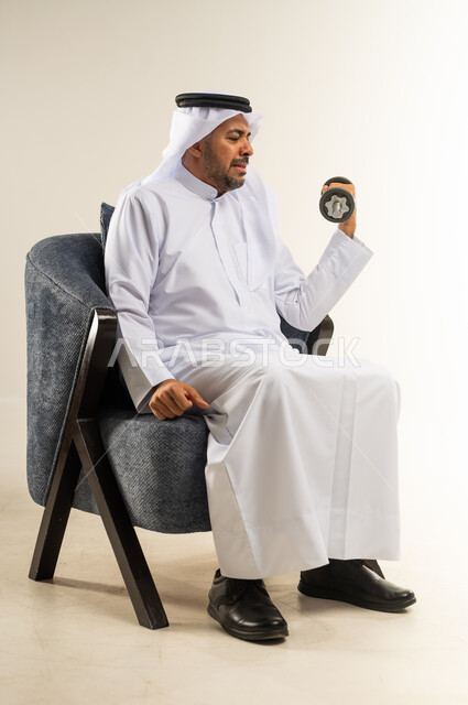 ممارسة التمارين الرياضية ورفع الأثقال مع الدمبل، بورتريه لسيد عربي خليجي كويتي يرتدي الثوب التقليدي يجلس على الكرسي المريح يحمل بيده الأوزان الحديدية الثقيلة، المحافظة على الصحة واللياقة الجسدية والرشاقة البدنية، خلفية بيضاء