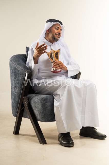 الاهتمام بالمظهر الخارجي، استخدام خشب العود في الأعياد والمناسبات، بورتريه لسيد عربي خليجي كويتي يرتدي الثوب التقليدي يجلس على كرسي مريح يقوم بالتطيب و التعطر من البخور( المبخرة ), العادات و التقاليد العريقة في الكويت, خلفية بيضاء