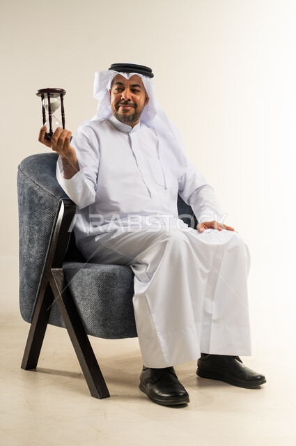 القيام بمراقبة توقيت الرمل, مفهوم إدارة الأعمال وتنظيم الجدول الزمني, بورتريه لسيد عربي خليجي كويتي يرتدي الثوب التقليدي يجلس على كرسي مريح يمسك بيده الساعة الرملية الزجاجية (البنكام) ينظر إليها بتعابير الفرح والسرور, إدراك قيمة اللحظات الثمينة, خلفية بيضاء