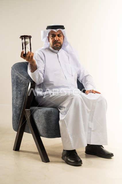 مفهوم إدارة الأعمال وتنظيم الجدول الزمني, بورتريه لسيد عربي خليجي كويتي يرتدي الثوب التقليدي يجلس على كرسي مريح يمسك بيده الساعة الرملية الزجاجية (البنكام) ينظر بتعابير الغضب والاستياء, إدراك قيمة اللحظات الثمينة, القيام بمراقبة توقيت الرمل, خلفية بيضاء