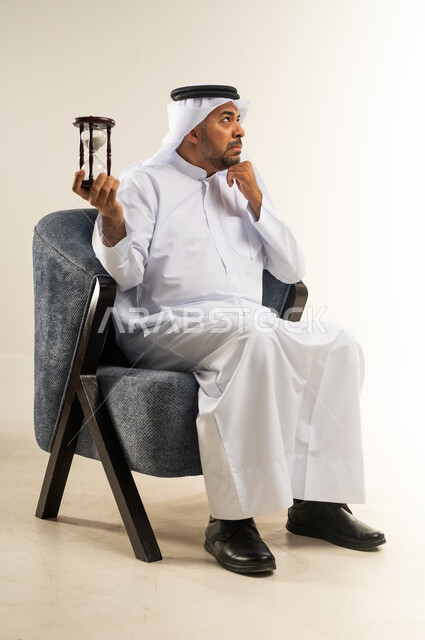 Realizing the value of precious moments, doing sand time monitoring, business management and schedule organization concept, portrait of a Kuwaiti Gulf Arab gentleman wearing traditional dress sitting on a comfortable chair holding in his hand a glass hourglass (bankam) putting his hand on his chin with thinking expression, white background
