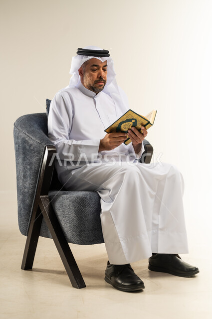 حفظ  كلام الله عز وجل، أداء العبادات والتقرب إلى الله تعالى، التعبد والتضرع بخشوع، بورتريه  لشاب عربي خليجي كويتي يرتدي الثوب التقليدي والغترة يجلس على كرسي مريح يقوم بتلاوة وترتيل القرآن الكريم, خلفية بيضاء
