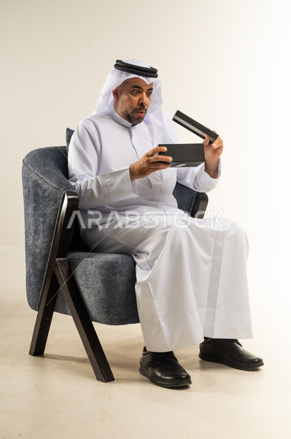 مفهوم المفاجأة والإهداء، إيماءات السعادة والدهشة بالحصول على هدية ثمينة، فن انتقاء الهدايا بعناية، بورتريه لشاب عربي خليجي كويتي يرتدي الثوب التقليدي والغترة يجلس على كرسي ويقوم بفتح العلبة بتعابير الصدمة، خلفية بيضاء