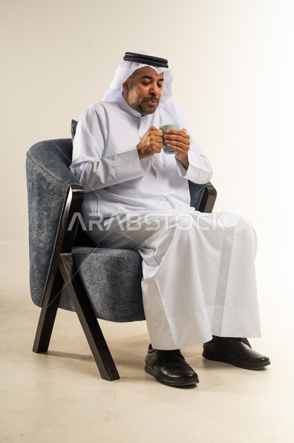 الاستمتاع وقضاء وقت ممتع بتناول المشروبات الساخنة، الحيوية والنشاط عند شرب الكافيين، بورتريه لرجل عربي خليجي كويتي يرتدي الثوب التقليدي والغترة يقوم بوضع يده على المشروب الساخن للاحساس بالدفء، خلفية بيضاء