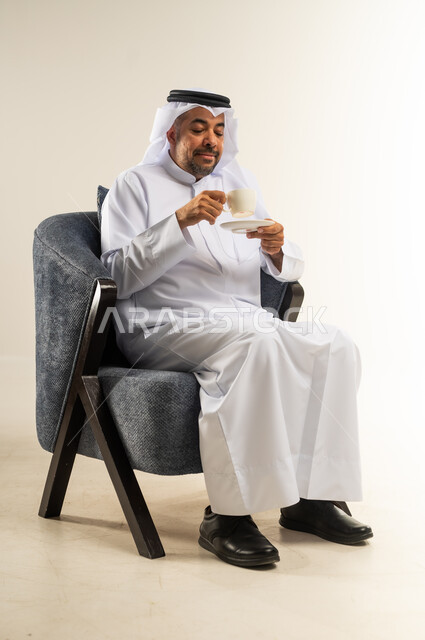 الاستمتاع والاحساس بالدفء عند تناول المشروبات الساخنة، الحيوية والنشاط عند شرب الكافيين، بورتريه لرجل عربي خليجي كويتي يرتدي الثوب التقليدي والغترة يحمل بيده كوب القهوة ويقوم بالنظر الى داخله بتعابير السرور، خلفية بيضاء