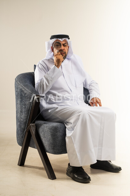 الشعور بالحيرة وعدم التأكد, مفهوم الاناقة والاهتمام بالمظهر الخارجي، الاحباط واليأس والعبوس، بورتريه لرجل عربي خليجي كويتي يرتدي الثوب التقليدي والغترة يجلس على كرسي يضع يده خلف ذقنه ينظر الى الكاميرا بتعابير الانزعاج، التخطيط لحل مشكلة ما، خلفية بيضاء