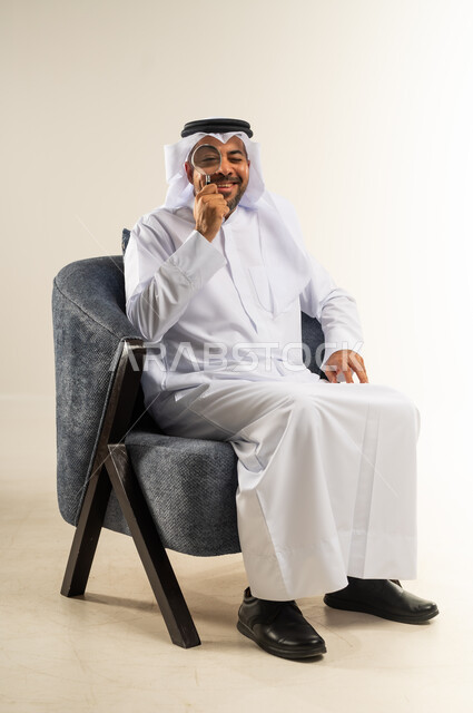 مفهوم الاناقة والاهتمام بالمظهر الخارجي، تعابير السعادة والسرور، بورتريه لرجل عربي خليجي كويتي مبتسم يرتدي الثوب التقليدي والغترة يجلس على كرسي يضع يده خلف ذقنه ينظر الى الكاميرا بإيماءات الفرح والثقة بالنفس، خلفية بيضاء