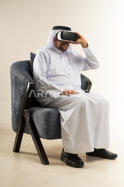 المتعة والتسلية  بالمشاهدة عبر نظارة الواقع الافتراضي, بورتريه من الجابب لرجل عربي خليجي كويتي يرتدي الثوب التقليدي والغترة يجلس على كرسي مريح يضع نظارات ثلاثية الأبعاد بتعابير الاندماج ومحاكاة العالم الخيالي, خلفية بيضاء