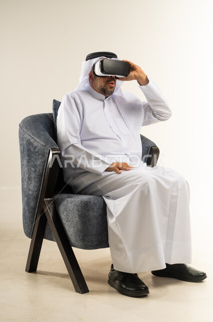 المتعة والتسلية  بالمشاهدة عبر نظارة الواقع الافتراضي, بورتريه من الجابب لرجل عربي خليجي كويتي يرتدي الثوب التقليدي والغترة يجلس على كرسي مريح يضع نظارات ثلاثية الأبعاد بتعابير الاندماج ومحاكاة العالم الخيالي, خلفية بيضاء