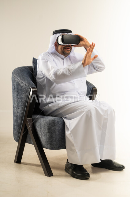 المتعة والتسلية  بالمشاهدة عبر نظارة الواقع الافتراضي, بورتريه لرجل عربي خليجي كويتي يرتدي الثوب التقليدي والغترة يجلس على اريكة مريحة يضع نظارات ثلاثية الأبعاد يقوم برفع وفتح كف يده للاعلى بتعابير الاندماج والتوقف عن فعل امر ما, خلفية بيضاء