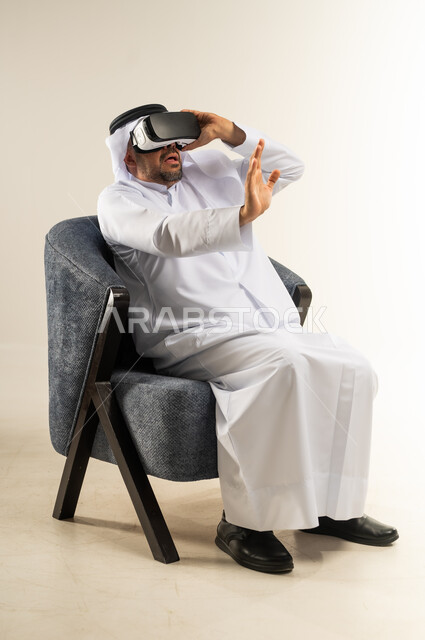 المتعة والتسلية  بالمشاهدة عبر نظارة الواقع الافتراضي, بورتريه لرجل عربي خليجي كويتي يرتدي الثوب التقليدي والغترة يجلس على اريكة مريحة يضع نظارات ثلاثية الأبعاد يقوم برفع وفتح كف يده للاعلى بتعابير الاندماج والتوقف عن فعل امر ما, خلفية بيضاء