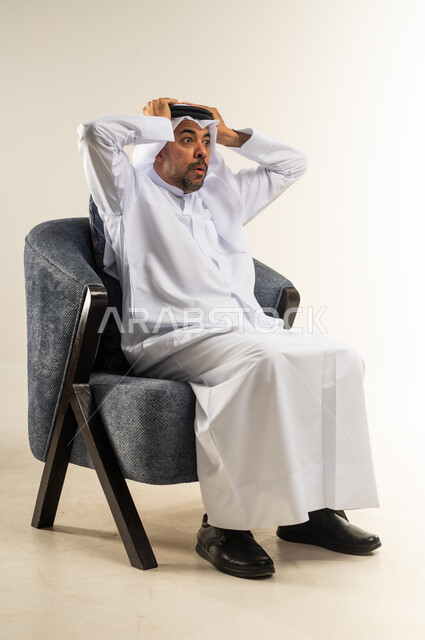 التفاجؤ من شيء ما، الشعور بالصدمة والذهول, بورتريه لرجل عربي خليجي كويتي يرتدي الثوب التقليدي والغترة يجلس على كرسي مريح ينظر الى الكاميرا ويضع يديه على رأسه بإيماءات التعجب والاندهاش, خلفية بيضاء