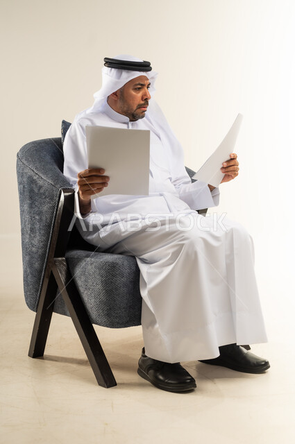 متابعة الخطط والمشاريع، قراءة الملاحظات والمعلومات، بورتريه من الجانب لرجل عربي خليجي كويتي يرتدي الثوب التقليدي والغترة يجلس على كرسي مريح يقوم بقراءة وتدقيق الاوراق  بتعابير التركيز والتمعن، مفهوم انجاز العمل، خلفية بيضاء