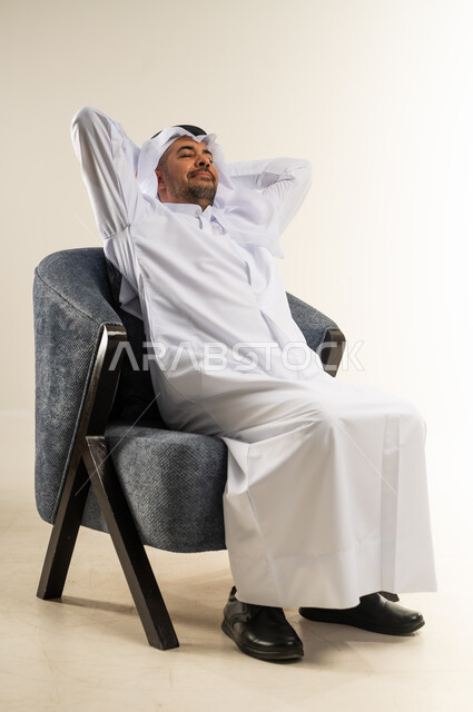 الشعور بالتعب والحاجة إلى الراحة، مفهوم الاسترخاء والاستمتاع بالأجواء الهادئة، بورتريه من الجانب لشاب عربي خليجي كويتي يرتدي االثوب التقليدي والغترة يجلس على كرسي مريح ويضع يديه خلف رأسه للنوم، خلفية بيضاء