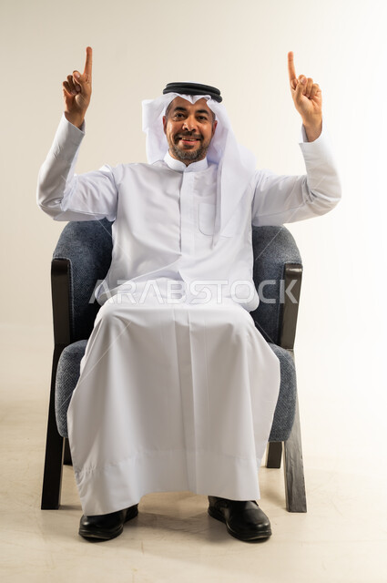 اشارة رقم واحد، النظر إلى الكاميرا بإيماءات الفرح والسرور، بورتريه لشاب عربي خليجي كويتي مبتسم يرتدي الثوب التقليدي والغترة يجلس على كرسي مريح يشير بإصبعيه السبابة للاعلى  إلى شيء محدد بتعابير هذا المكان هنا، خلفية بيضاء .