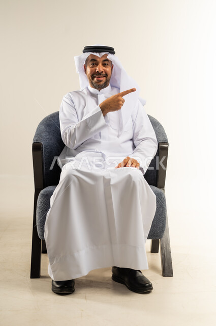 النظر إلى الكاميرا بإيماءات الفرح والسرور، بورتريه لشاب عربي خليجي كويتي مبتسم يرتدي الثوب التقليدي والغترة يجلس على كرسي مريح يشير بإصبعه السبابة جانبا  إلى شيء محدد بتعابير هذا المكان هنا، خلفية بيضاء .