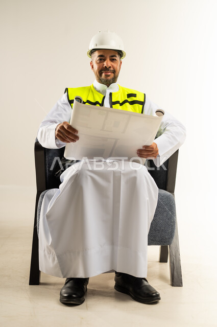العمل في القطاع الهندسي، بورتريه لرجل عربي خليجي كويتي يرتدي الثوب التقليدي مع سترة الحماية والغترة يجلس على كرسي مريح يمسك مخططات الإعمار بيديه، النظر الى الكاميرا بتعابير الفخر والسرور، إدارة ومتابعة المشروع, مفهوم الهندسة والانشاءات المعمارية, خلفية بيضاء