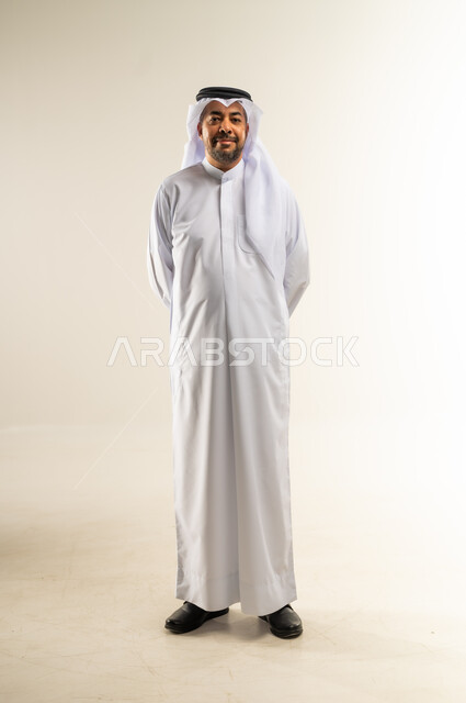 الاهتمام بالأناقة والمظهر الخارجي، بورتريه لرجل عربي خليجي كويتي يرتدي الثوب التقليدي والغترة يقف باستقامة يضع يديه خلف ظهرة بإيماءات السرور والثقة في النفس، مفهوم الوسامة والرجولة والقوة، الجسم الكامل بالطول، خلفية بيضاء