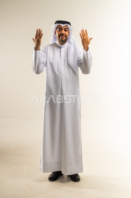 مفهوم الاناقة والاهتمام بالمظهر الخارجي، عرض ظاهر الكف والنظر الى الكاميرا بتعابير الفرح والسرور، بورتريه لرجل عربي خليجي كويتي مبتسم يرتدي الثوب التقليدي  والغترة يقف و يرفع كفيه مفتوحة للاعلى، خلفية بيضاء