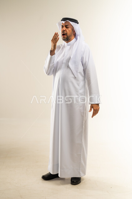 الشعور بالتعب والحاجة الى النوم والراحة, الإحساس بالكسل والخمول و الإرهاق، بورتريه  لرجل عربي خليجي كويتي يرتدي الثوب التقليدي والغترة يضع يده على فمه بإيماءات التثاؤب والنعاس، صورة الجسم بالطول الكامل, خلفية بيضاء