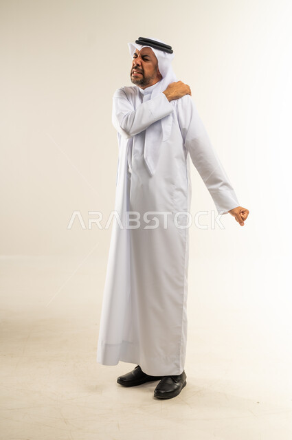الأوجاع والآلام العصبية والعضلية، طلب المساعده والحاجه للعلاج، التهاب العضلات والمفاصل، بورتريه من الجانب لرجل عربي خليجي كويتي يرتدي الثوب التقليدي والغترة يضع يده على كتفه بايماءات الألم والتعب والأرهاق، صورة الجسم الطول الكامل, خلفية بيضاء
