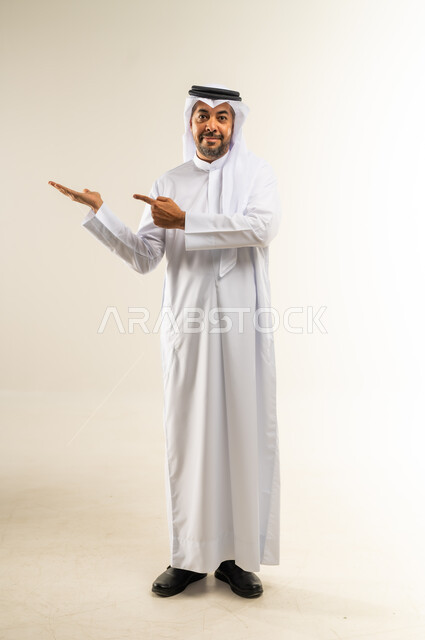 النظر إلى الكاميرا بإيماءات الفرح والسرور، بورتريه لشاب عربي خليجي كويتي مبتسم يرتدي الثوب التقليدي والغترة يقف ويشير بإصبعيه السبابة جانبا  إلى شيء محدد بتعابير هذا المكان هنا، الجسم الكامل بالطول، خلفية بيضاء .