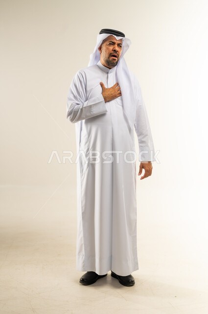 Feeling an increase in heart rate, the need to go to the doctor to conduct the necessary tests, heart disorders and diseases, a portrait of a Kuwaiti Gulf Arab man wearing a traditional dress and a ghutra, putting his hand on his chest, feeling a tingling and pain in his heart, a full-length body image, white background