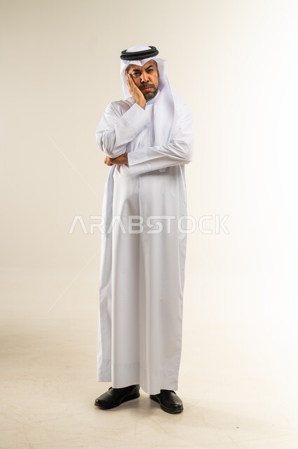 ايماءات الشعور بالإحباط واليأس وخسارة شيء ما، بورتريه لرجل عربي خليجي كويتي يرتدي الثوب التقليدي والغترة يقف باستقامة ويضع يده على خده بتعابير الاكتئاب والزعل وعدم الرضا، الجسم الكامل بالطول، خلفية بيضاء