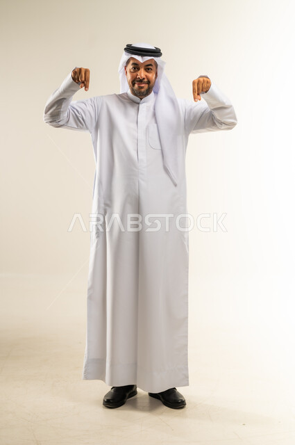 Confirming something is happening right now, gestures indicating this place (here), specifying a specific location, portrait of a smiling Kuwaiti Gulf Arab man wearing traditional thobe and ghutra pointing index fingers downwards, looking at the camera with gestures of pleasure, full body length, white background