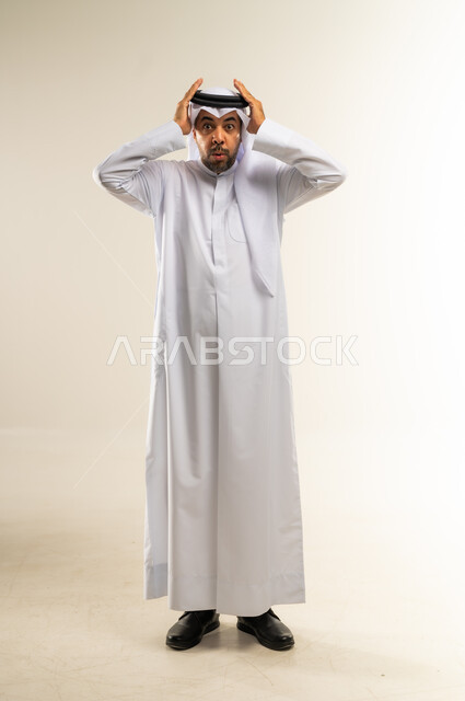 التفاجؤ من سماع خبر سيء، الشعور بالدهشة والصدمة والذهول, بورتريه لشاب عربي خليجي كويتي يرتدي الثوب والغترة ينظر الى الكاميرا ينظر الى الكاميرا يضع يديه على رأسه بإيماءات الخوف والذعر, صورة الجسم بالطول الكامل, خلفية بيضاء
