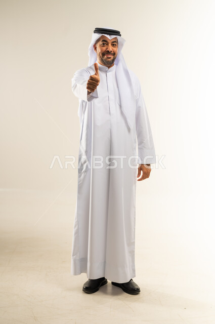 تعابير القبول والفرح والسرور، الاهتمام بالأناقة والمظهر الخارجي، بورتريه لرجل عربي خليجي كويتي  يرتدي الثوب التقليدي والغترة ينظر الى الكاميرا ويرفع إصبع الابهام للاعلى بإيماءات الإعجاب والرضا والموافقة، الطول الكامل للجسم، خلفية بيضاء