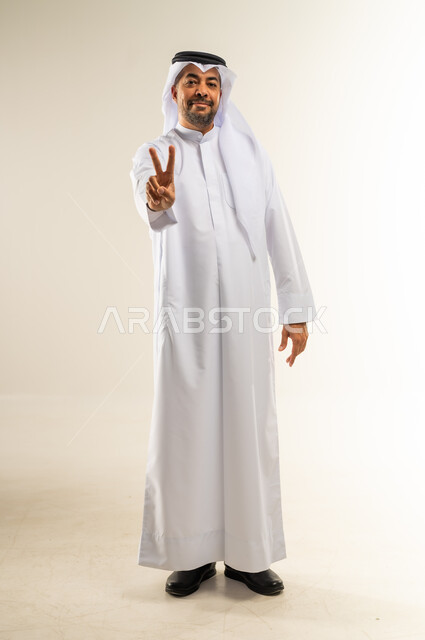 رفع علامة النصر والسلام، إشارة الفوز والانتصار، تحقيق الأهداف والطموحات، إنجاز المهام المحددة، إشارات وحركات لغة الجسد، بورتريه لرجل عربي خليجي كويتي مبتسم يرتدي الثوب التقليدي والغترة ينظر للكاميرا بإيماءات السعادة والسرور، الجسم بالطول الكامل، خلفية بيضاء