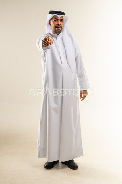 توجيه أصابع الاتهام إلى شخص معين، الإشارة باصبع السبابة إلى الكاميرا بشكل مباشر، التهديد والتوعد بتنفيذ شيء ما، إيماءات التحذير والترهيب، بورتريه لرجل عربي خليجي كويتي يرتدي الثوب التقليدي والغتره يشير إلى مكان ما محدد، الجسم بالطول الكامل، خلفية بيضاء