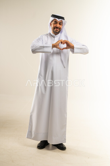 إظهار الحب للآخرين، التعبير عن المحبة والمودة، الاهتمام بالمظهر الخارجي، مفهوم الأناقة والوسامة، النظر إلى الكاميرا بإيماءات البشاشة، بورتريه لرجل عربي خليجي كويتي مبتسم يرتدي الثوب التقليدي والغتره يقوم بصنع شكل قلب بيديه، الجسم بالطول الكامل، خلفية بيضاء