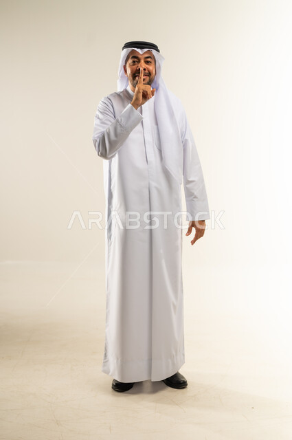الأمر بالصمت التام، وضع اصبع السبابة أمام الفم بإيماءات الجدية، طلب التوقف عن الكلام والصمت، النهي والزجر والانصات، بورتريه لرجل عربي خليجي كويتي يرتدي الثوب التقليدي والغتره ينظر إلى الكاميرا بإيماءات البشاشة، الجسم بالطول الكامل، خلفية بيضاء