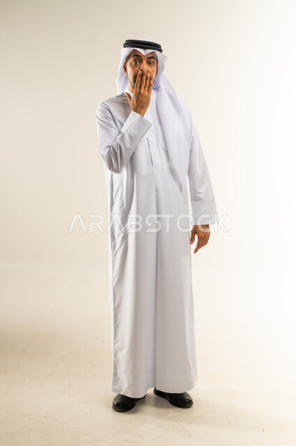التعرض لصدمة وعدم التصديق، النظر إلى الكاميرا بإيماءات الدهشة والمفاجأة، خيبة الأمل والمشاعر السلبية، بورتريه لرجل عربي خليجي كويتي يرتدي الثوب التقليدي والغتره يضع يده أمام فمه، الاهتمام بالمظهر الخارجي، الجسم بالطول الكامل، خلفية بيضاء