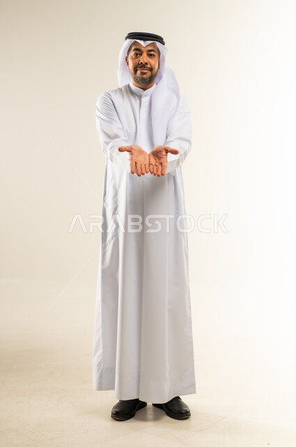 تقديم الضيافة أو إعطاء شيء ما، فتح الكفين ومدهما محو الأمام، مفهوم التقدير والاحترام، بورتريه لرجل عربي خليجي كويتي مبتسم يرتدي الثوب التقليدي والغتره ينظر إلى الكاميرا بإيماءات السعادة والسرور، الأناقة والوسامة والرجولة، الجسم بالطول الكامل، خلفية بيضاء