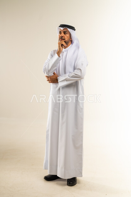 التفكير بعمق وشرود الذهن، محاولة إيجاد حل للمشكلات العصبة، القلق والحيرة في اتخاذ القرار الصحيح، بورتريه لرجل عربي خليجي قطري مبتسم يرتدي الثوب التقليدي والغتره يضع يده أسفل ذقنه وينظر إلى مكان ما بعيد بإيماءات الاندماج، الجسم بالطول الكامل، خلفية بيضاء